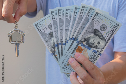 Asian man holding one hundred dollar bills and home shape key chain. Property investment and mortgage finance concept