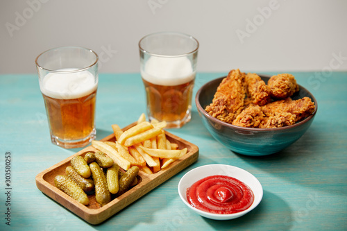 delicious chicken nuggets, ketchup, french fries and gherkins near glasses of beer on turquoise wooden table isolated on grey