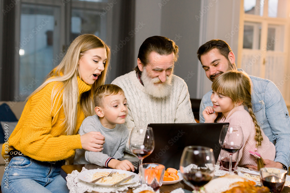 Happy smiling family looking film or making call via internet using laptop, sitting at the festive tabe at home, celebrating dinner together. Thanksgiving day concept
