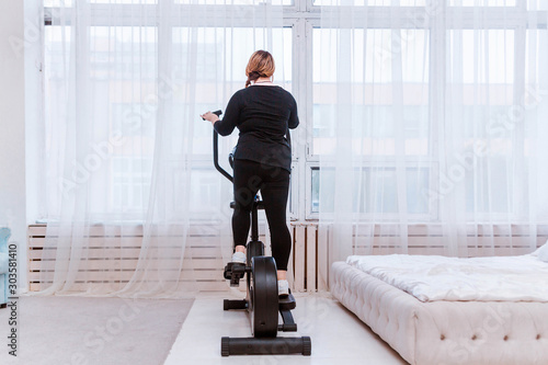 Model - a fat woman, trying to lose weight and at home is engaged in an elliptical trainer photo