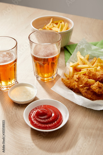 glasses of beer, chicken nuggets with french fries, ketchup and mayonnaise on wooden table on grey background