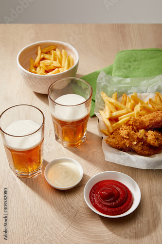 glasses of beer, chicken nuggets with french fries, ketchup and mayonnaise on wooden table on grey background