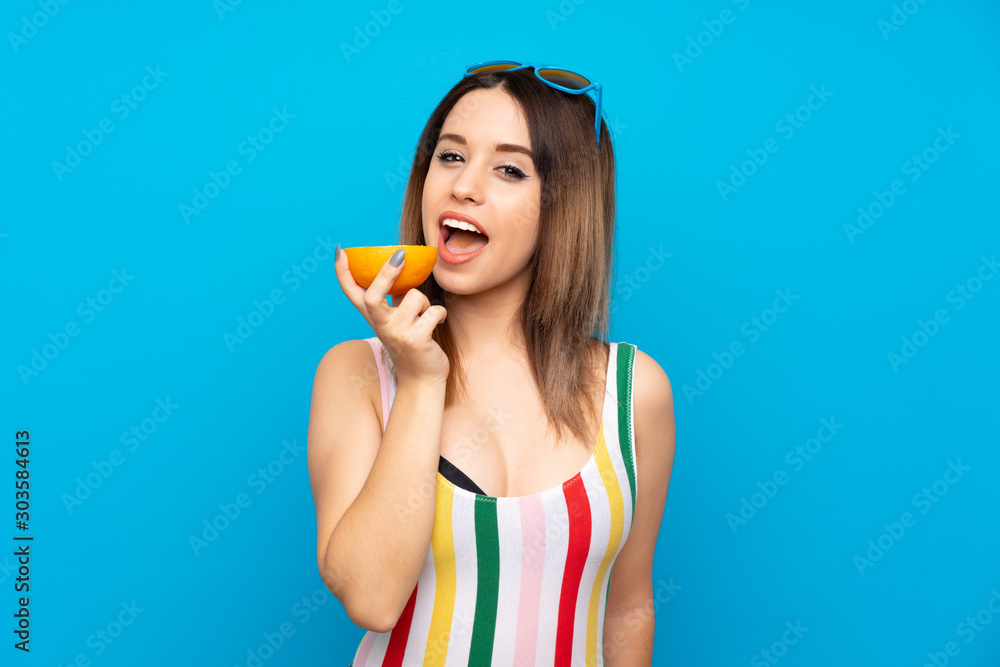 Young woman in summer holidays over blue background holding an orange Young woman in summer holidays over blue background holding an orange