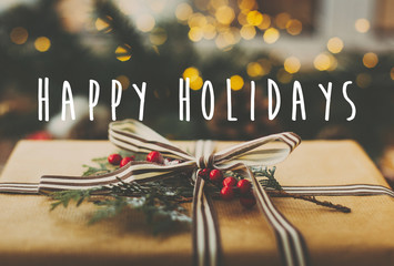 Happy Holidays text sign on stylish rustic christmas gift and golden lights, pine branches, cones on rural wooden table. Season's greeting card