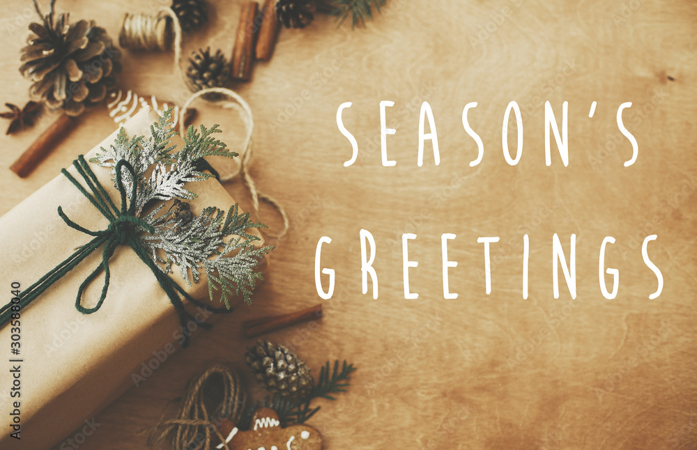 Season's greetings text sign on stylish rustic christmas gifts box with cedar branch on rural table with pine cones, gingerbread cookies, cotton, cinnamon. Season's greeting card - obrazy, fototapety, plakaty 