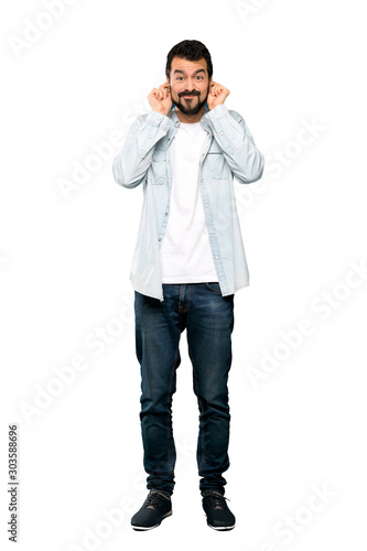 Full-length shot of Handsome man with beard frustrated and covering ears over isolated white background © luismolinero
