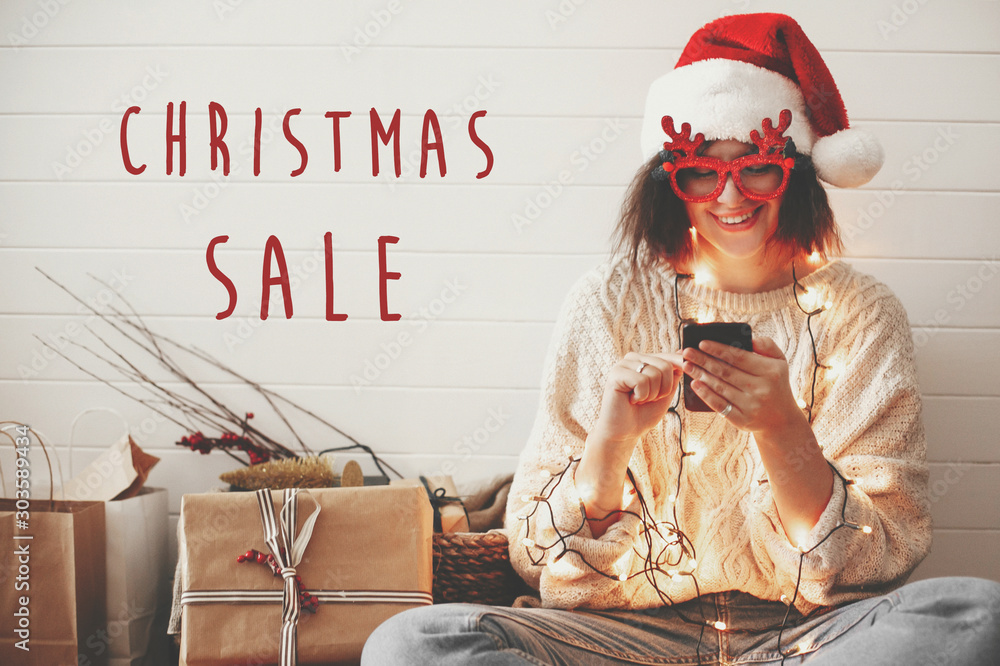 Φωτογραφία Christmas Sale text sign on stylish happy girl in santa hat looking at phone screen in festive christmas lights on background of  presents