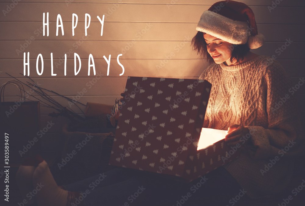 Φωτογραφία Happy Holidays text sign on stylish happy girl in santa hat opening christmas gift box with magic light in dark room