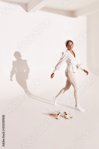 full-length photo of dark-skinned ballerina on white background with shadow on it, she wearing white shirt, beige bodysuit, white tights she stands near her pointes and looks down