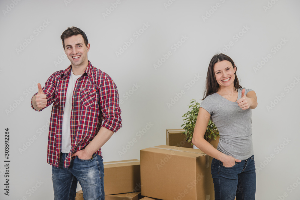 Happy man and woman standing near stack of carton boxes in their new apartment, giving thumbs up, putting one hand into pocket, smiling. Happy man and woman standing near stack of carton boxes in their new apartment, giving thumbs up, putting one hand into pocket, smiling.