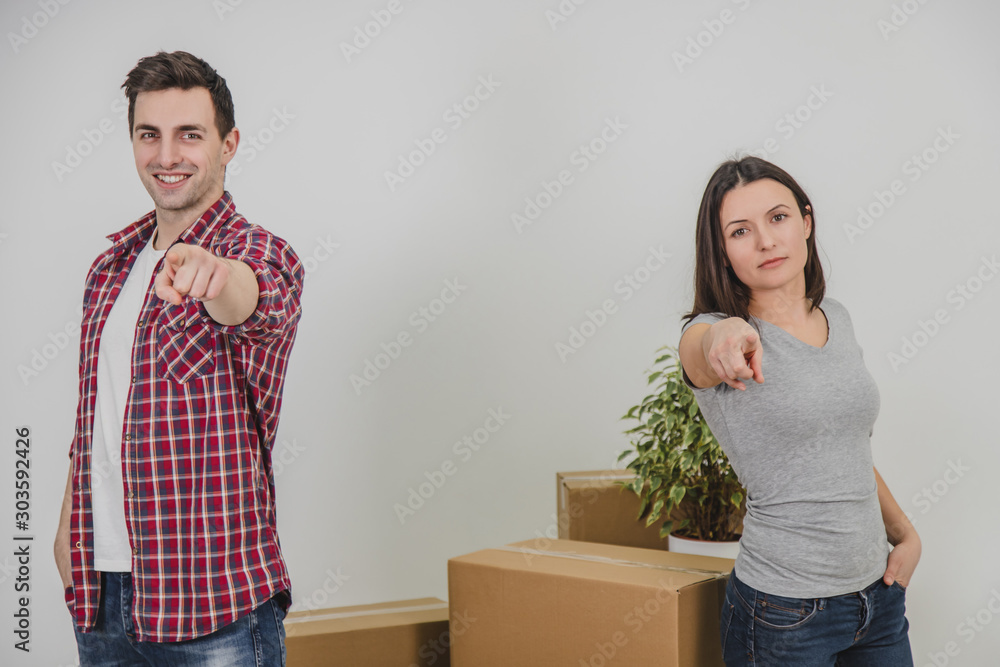 Happy man and woman standing side by side near stack of carton boxes in their new apartment, pointing their fingers at the camera, smiling. Happy man and woman standing side by side near stack of carton boxes in their new apartment, pointing their fingers at the camera, smiling.