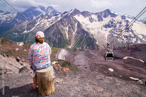 A child girl sits on a stump standing on the observation deck in the summer and looks at the mountain scenery in the mountains of the Caucasus.