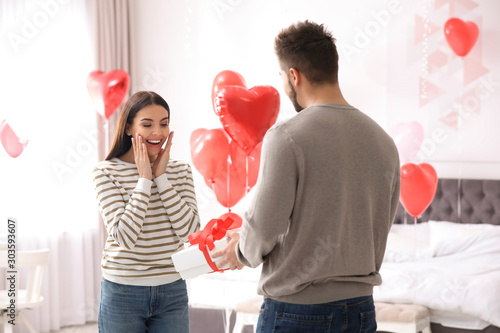 Young man presenting gift to his girlfriend in bedroom decorated with heart shaped balloons. Valentine's day celebration