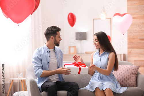 Young man presenting gift to his girlfriend in living room decorated with heart shaped balloons. Valentine's day celebration