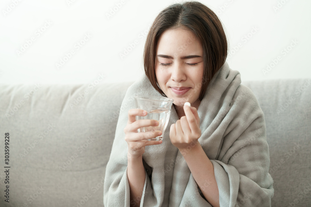 Asian Cute of girl having headache and high temperature from illness holding pill of medicine and water sitting on sofa at home, Health and illness concepts Asian Cute of girl having headache and high temperature from illness holding pill of medicine and water sitting on sofa at home, Health and illness concepts
