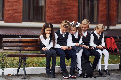 Group of kids in school uniform sits on the bench outdoors together near education building