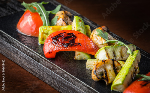 Grilled vegetables on wooden board on dark background. Grilled peppers, tomatoes, mushrooms and green zucchini decorated with arugula. Сloseup