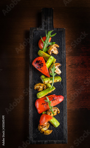 Grilled vegetables on wooden board on dark background. Grilled peppers, tomatoes, mushrooms and green zucchini decorated with arugula. Horizontal view from above