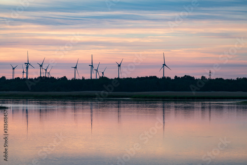 Evening sunlight on coast, pink and golden clouds and wind turbine. Sky reflection on water.  Wind generator for electricity, alternative energy source. Windmill for electric power production. © Artenex