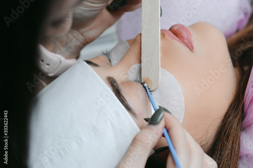 Eyelash Extension Procedure. Close up view of beautiful Woman with Long Eyelashes. Stylist holding tweezers, tongs and making lengthening lashes for girl in a beauty salon. Beauty Concept.
