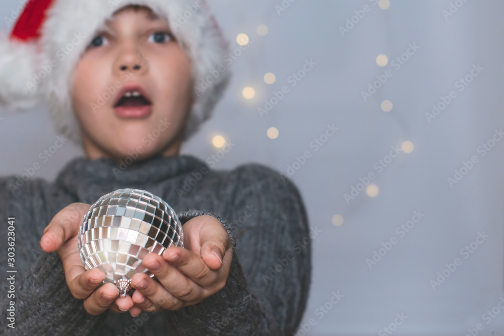 Naklejka premium A surprised 9-year-old boy in a Santa hat is holding a shiny disco ball. Toning. New year context. Emotions.