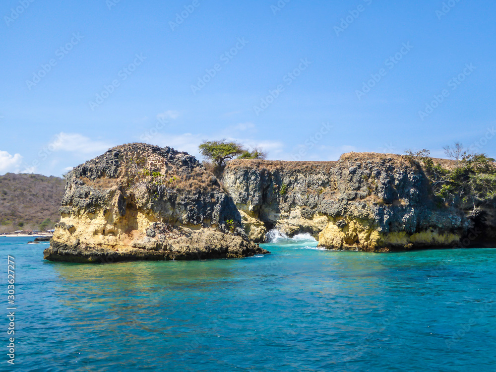 A cliff formation emerging from the calm sea. The water has many shades of turquoise. Sunny and cloudless day. Unspoiled and raw beaches of Indonesia. Hidden gem.