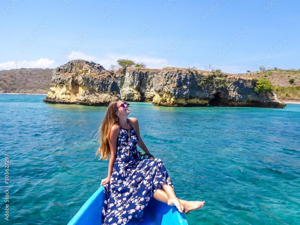 A girl in a maxi dress sitting on a blue boat and enjoying the breeze in her hair. In the back there is a cliff formation emerging from the calm sea. The water has many shades of turquoise. Happiness