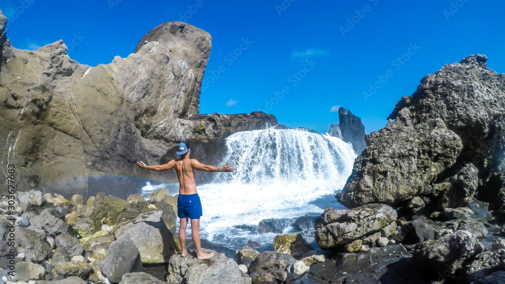 Young man standing in front of the Wave waterfall on Pantai Nambung, Lombok, Indonesia.The water crushes on the rocks on shore, breaching through them, creating a temporary waterfall. Great adventure Young man standing in front of the Wave waterfall on Pantai Nambung, Lombok, Indonesia.The water crushes on the rocks on shore, breaching through them, creating a temporary waterfall. Great adventure