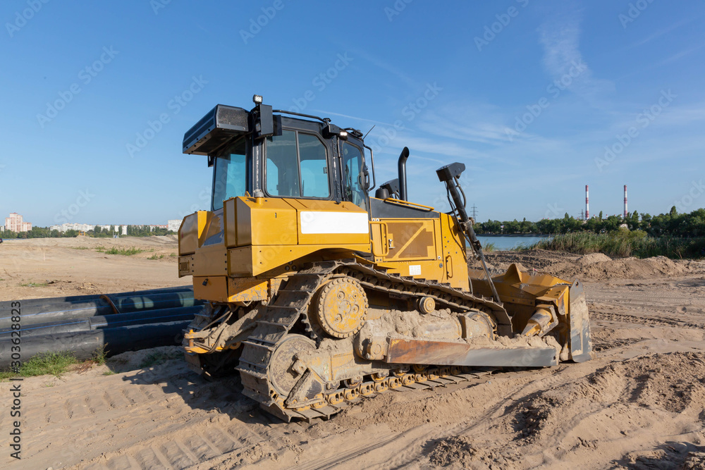 A crawler, yellow, black-roofed bulldozer stands on the sandy surface of the dam under a blue sky. A crawler, yellow, black-roofed bulldozer stands on the sandy surface of the dam under a blue sky.