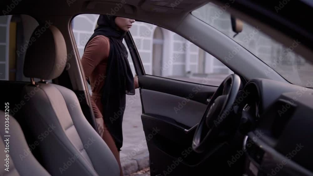 Young beautiful muslim girl in black hijab open the door and sitting in car, watching at mirror, fixing it. Insert the keys - starts the engine, putting on the safe belt - ready to drive. Side footage Young beautiful muslim girl in black hijab open the door and sitting in car, watching at mirror, fixing it. Insert the keys - starts the engine, putting on the safe belt - ready to drive. Side footage