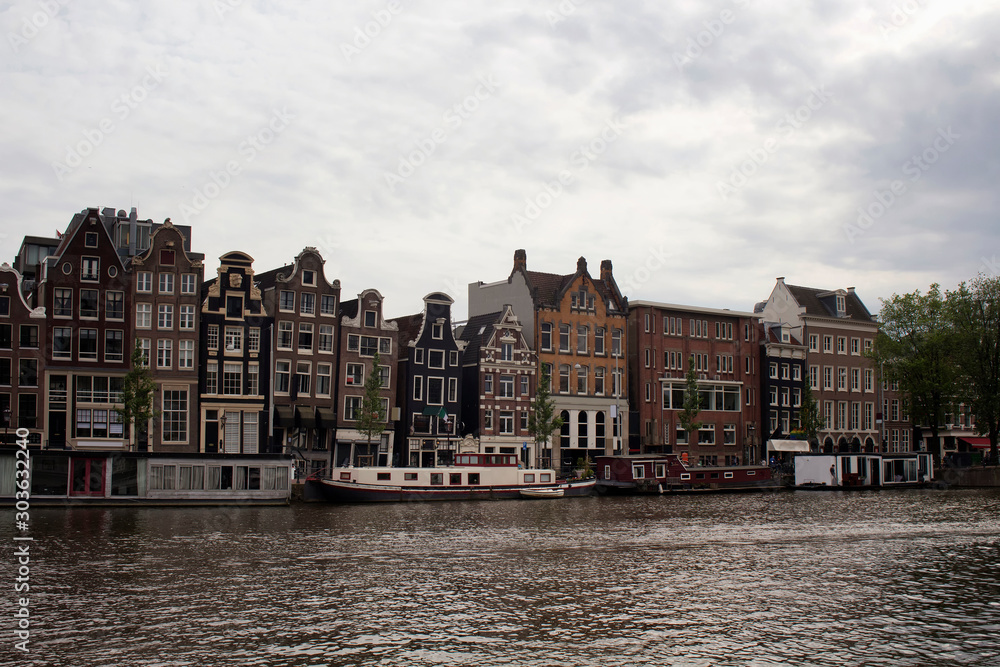 View of Amstel river, canal boat houses and historical, traditional and typical buildings reflecting Dutch architectural style in Amsterdam. It is a summer day with cloudy sky.