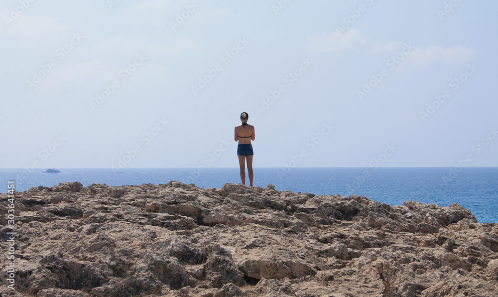 Girl in denim shorts on a rock overlooking the sea. The view from the back.