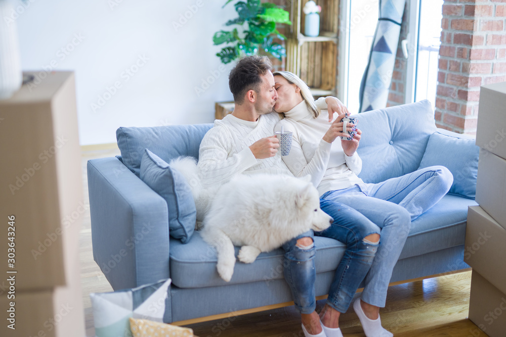 Young beautiful couple with dog sitting on the sofa drinking coffee at new home around cardboard boxes Young beautiful couple with dog sitting on the sofa drinking coffee at new home around cardboard boxes