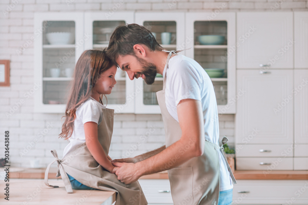 Fototapeta premium daughter sitting on the table and dad standing on the kitchen and looking at each other, side view