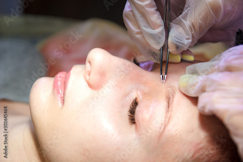 A beautician in a beauty salon plucks eyebrows on a client's face with tweezers