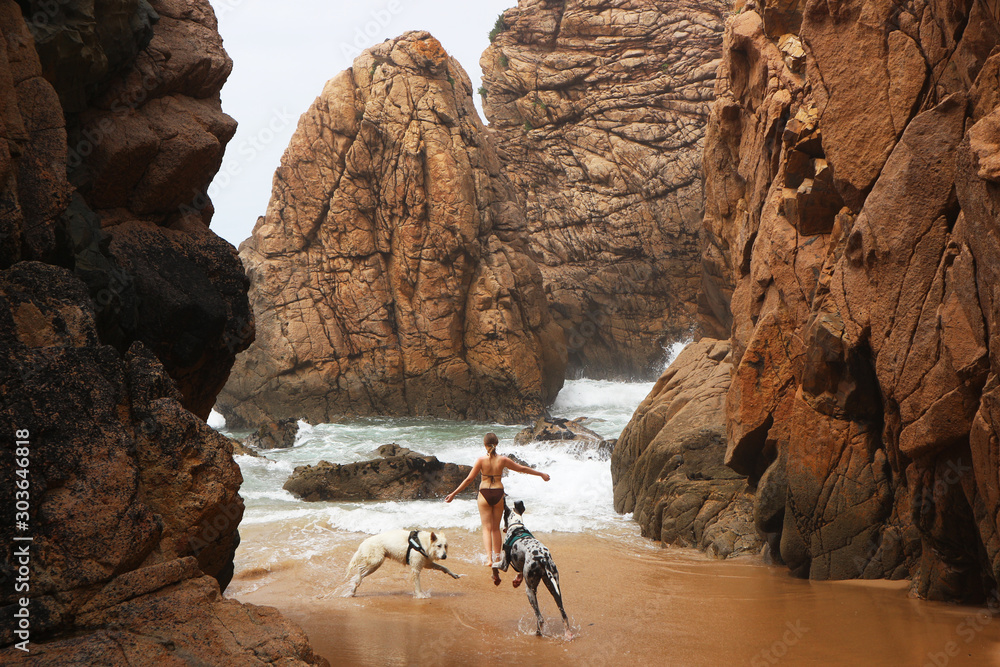 Young woman or sporty active girl playing with two dogs outside on sandy rocky beach near the ocean. Praia da ursa or Ursa Beach. Near the Cabo da Roca. Sintra-Cascais Natural Park.