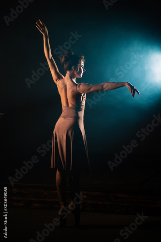 Ballerina is practicing her moves on dark stage. Young girl dancing in beige dress, spinning around and smiling. Gracefulness and tenderness in every movement.