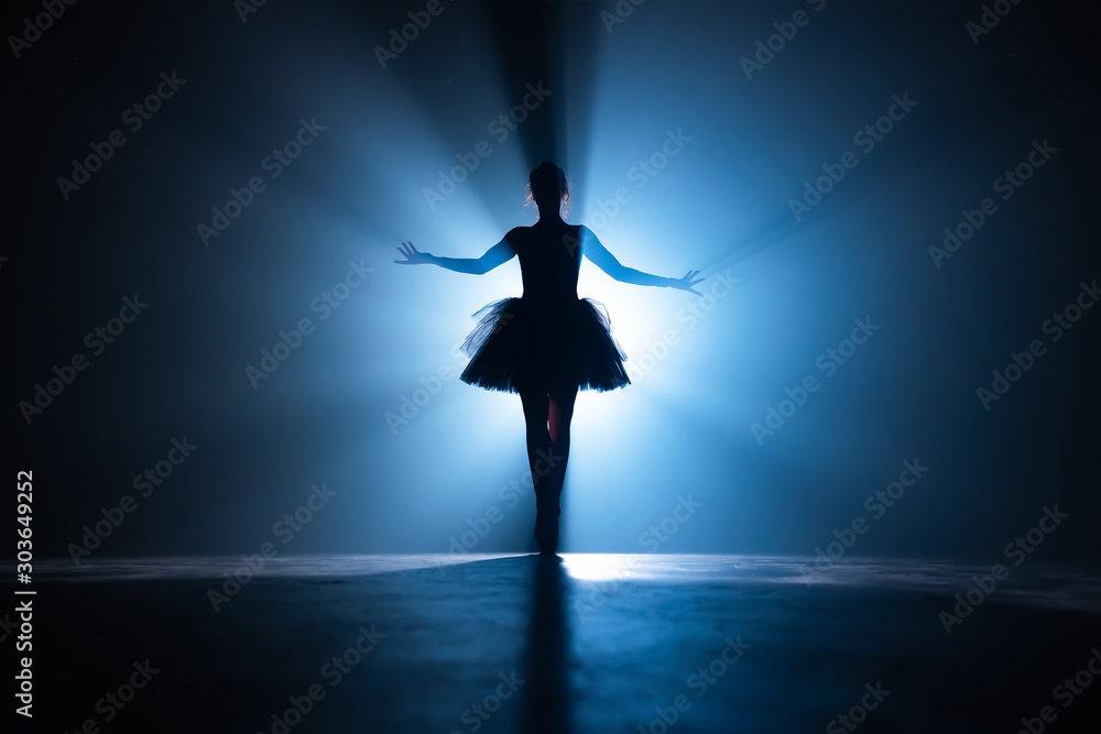 Young beautiful ballerina on smoke stage dancing modern ballet. Performs smooth movements with hands against spotlight background. Woman in black tutu costume on scene.
