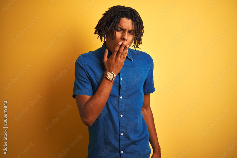 Afro man with dreadlocks wearing casual denim shirt standing over isolated yellow background bored yawning tired covering mouth with hand. Restless and sleepiness. Afro man with dreadlocks wearing casual denim shirt standing over isolated yellow background bored yawning tired covering mouth with hand. Restless and sleepiness.