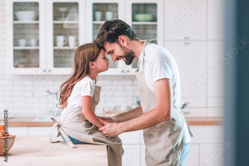 side view of father and girl wearing aprons looking at each other on the kitchen photo