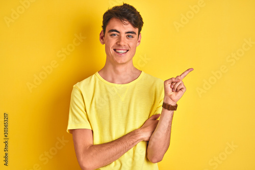 Teenager boy wearing yellow t-shirt over isolated background with a big smile on face, pointing with hand and finger to the side looking at the camera.