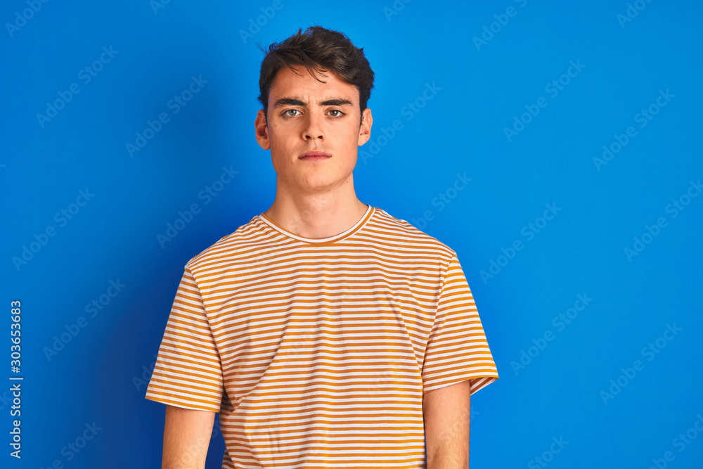 Teenager boy wearing casual t-shirt standing over blue isolated background with serious expression on face. Simple and natural looking at the camera. Teenager boy wearing casual t-shirt standing over blue isolated background with serious expression on face. Simple and natural looking at the camera.
