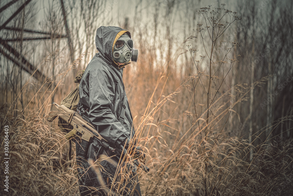 Soldier with rifle and bag in raincoat and gas mask is walking away through dried grass.