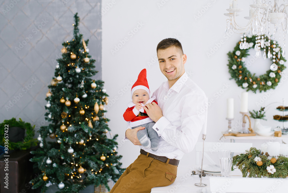 Φωτογραφία A young father holds a young son in a little Santa costume on the top of a Christmas tree in the living room