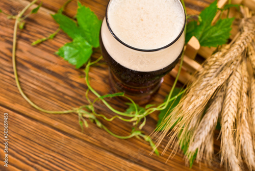Glass of dark beer with natural green hop leaves and dry wheat ears on a wooden background.