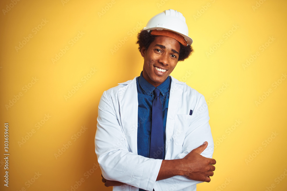 Afro american engineer man wearing white coat and helmet over isolated yellow background happy face smiling with crossed arms looking at the camera. Positive person. Afro american engineer man wearing white coat and helmet over isolated yellow background happy face smiling with crossed arms looking at the camera. Positive person.