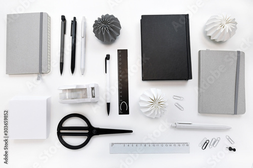 High angle top view of white desk with various black, gray and white neatly arranged office supplies like scissors, pens, pencils and notebooks