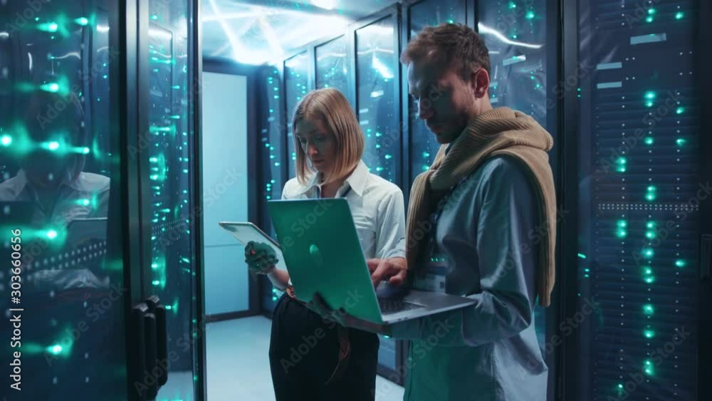 Couple of server engineers cooperating in high tech data center. Male and female technicians updating hardware inspecting system performance in server room. Couple of server engineers cooperating in high tech data center. Male and female technicians updating hardware inspecting system performance in server room.