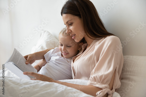 Young mommy lying in bed with daughter, reading book.