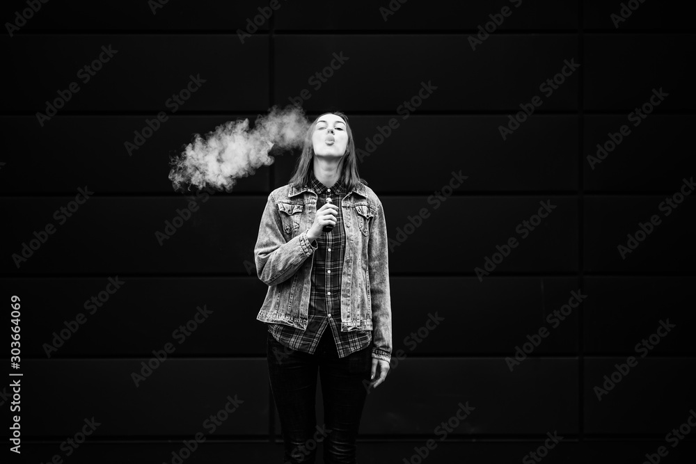 Vaping teenager. Young pretty white girl in casual clothes smoking an electronic cigarette opposite dark modern backgroun on the street in the autumn. Bad habit. Vape activity. Black and white. Vaping teenager. Young pretty white girl in casual clothes smoking an electronic cigarette opposite dark modern backgroun on the street in the autumn. Bad habit. Vape activity. Black and white.